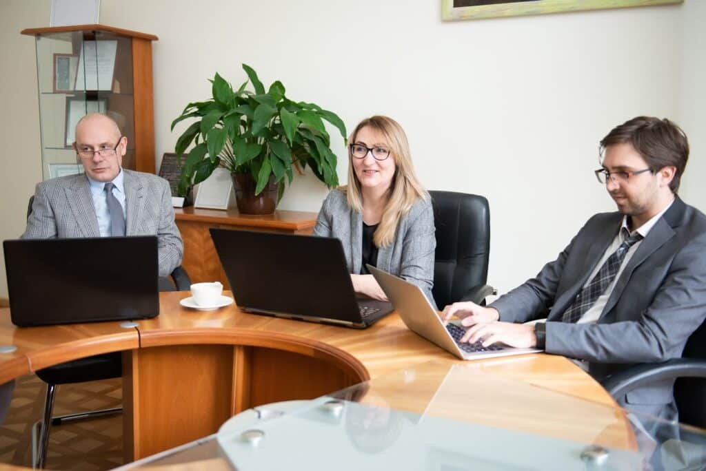 Three people in business attire sit around a wooden table, each working on a laptop with financial statements. A potted plant is in the background. One person smiles while others focus on their screens. A cup of coffee sits on the table.