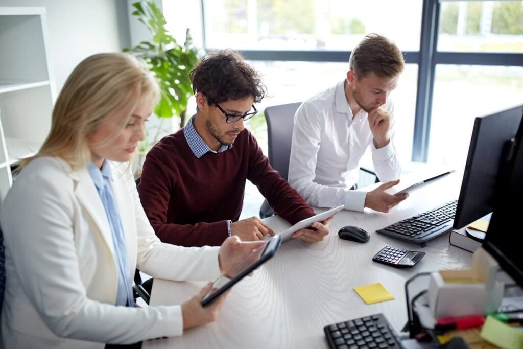 Three people sit at a desk in an office, focused on financial statements tablets. They are surrounded by computers, office supplies, and a large window. The atmosphere appears collaborative and professional.