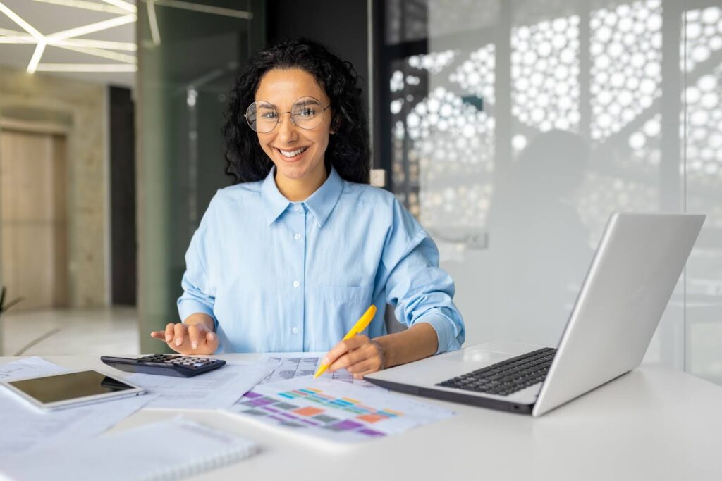 A person with curly hair and glasses is smiling while sitting at a desk with working on financial statements. They hold a yellow pencil and have a laptop, calculator, and papers with colorful charts in front of them. The setting is a bright office with modern decor.