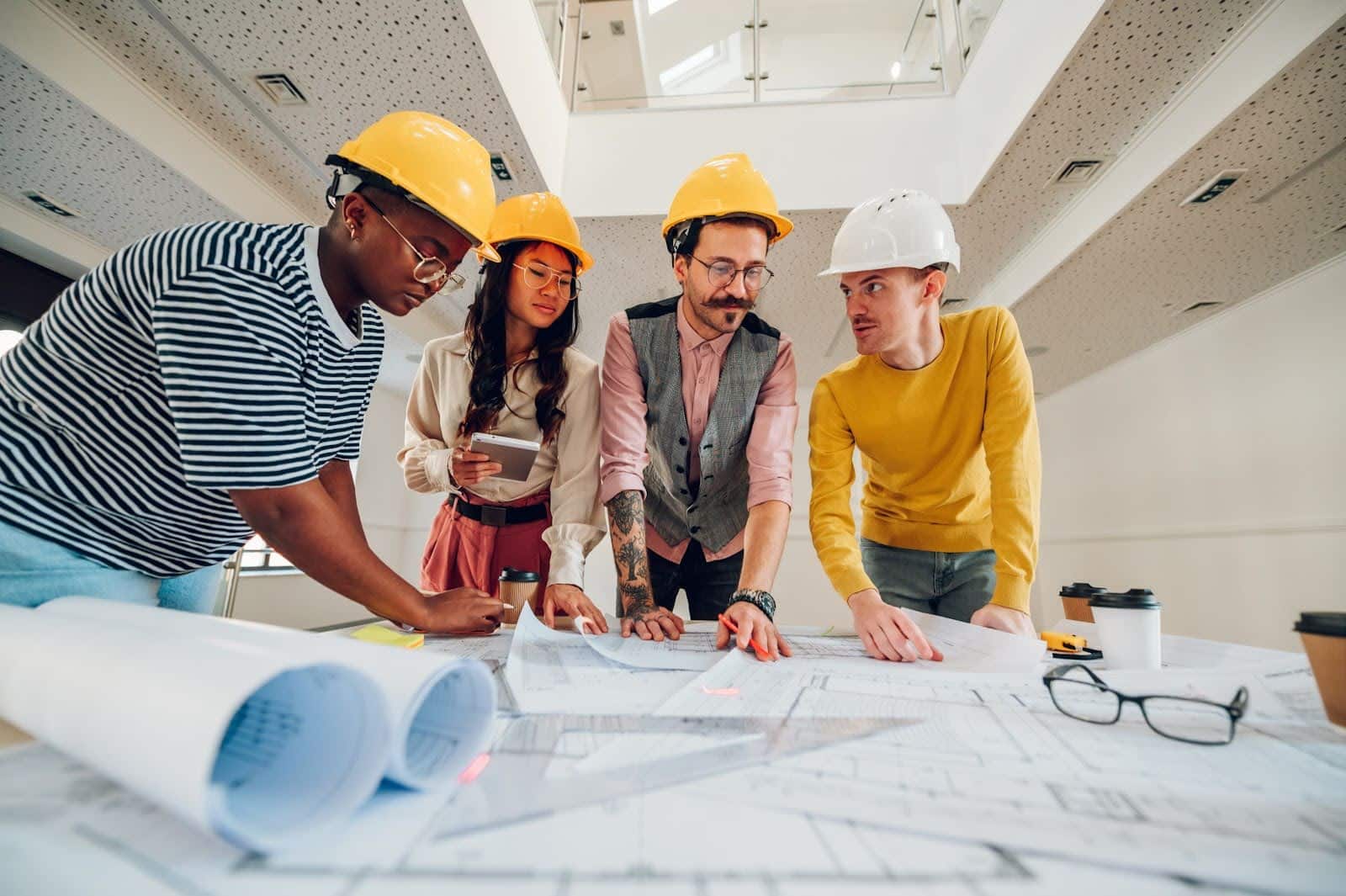 Four people in hard hats gather around a table with architectural blueprints. They appear to be discussing the plans and cash flow management and are in a modern office setting. Coffee cups and rolled-up plans are also visible on the table.