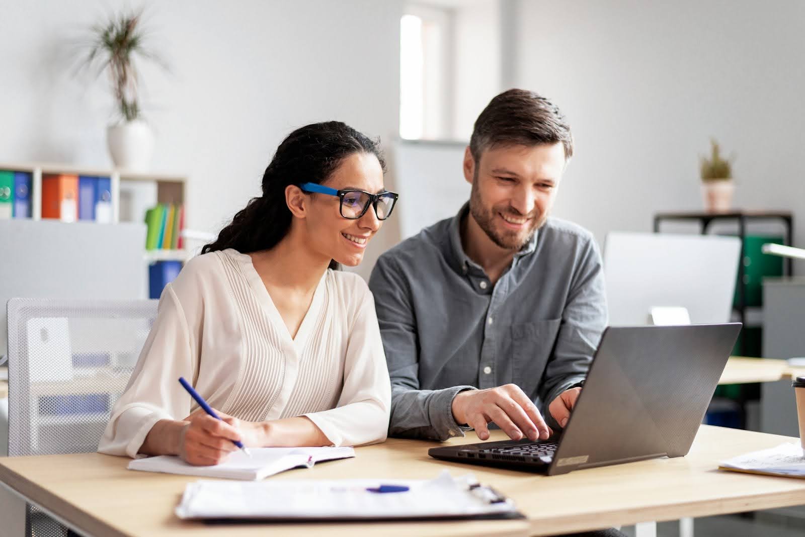 A woman and a man sit at a desk in an office, smiling and looking at financial close automation on a laptop. The woman is holding a pen and notebook. The office is bright, with shelves and plants in the background.