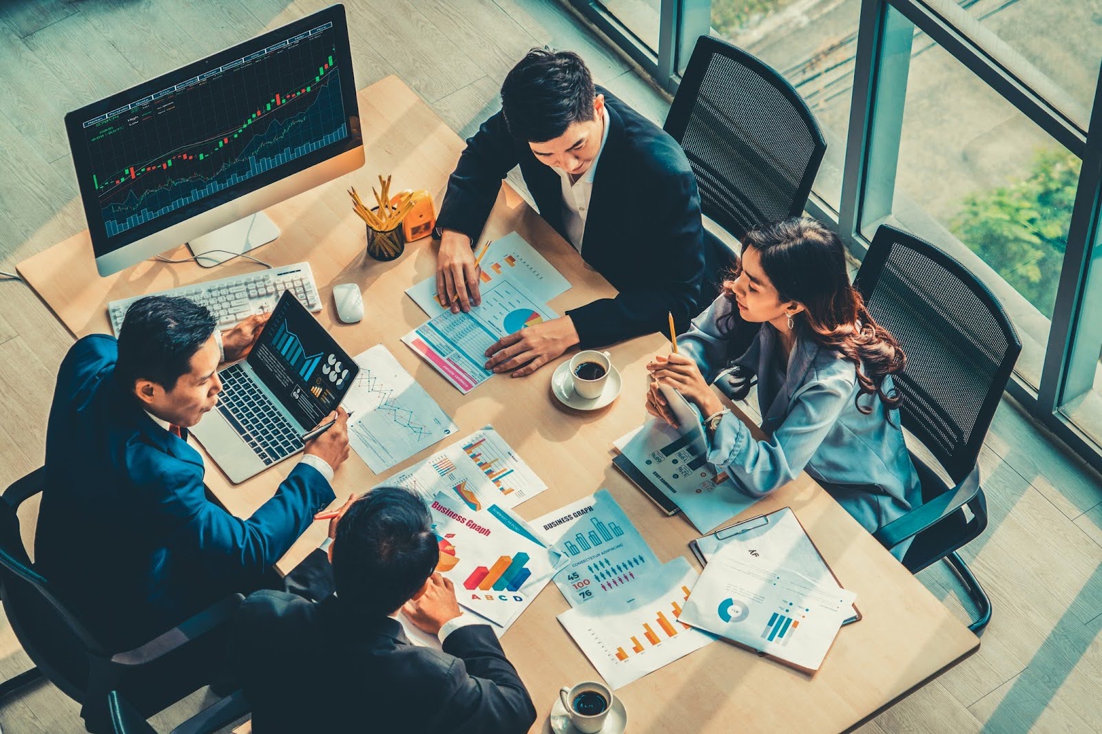 A group of four business professionals in formal attire sit around a table in an office, meticulously reviewing charts and graphs on papers and a laptop. A large monitor is visible in the background, displaying a financial graph that underscores the importance of compliance automation in their strategy.