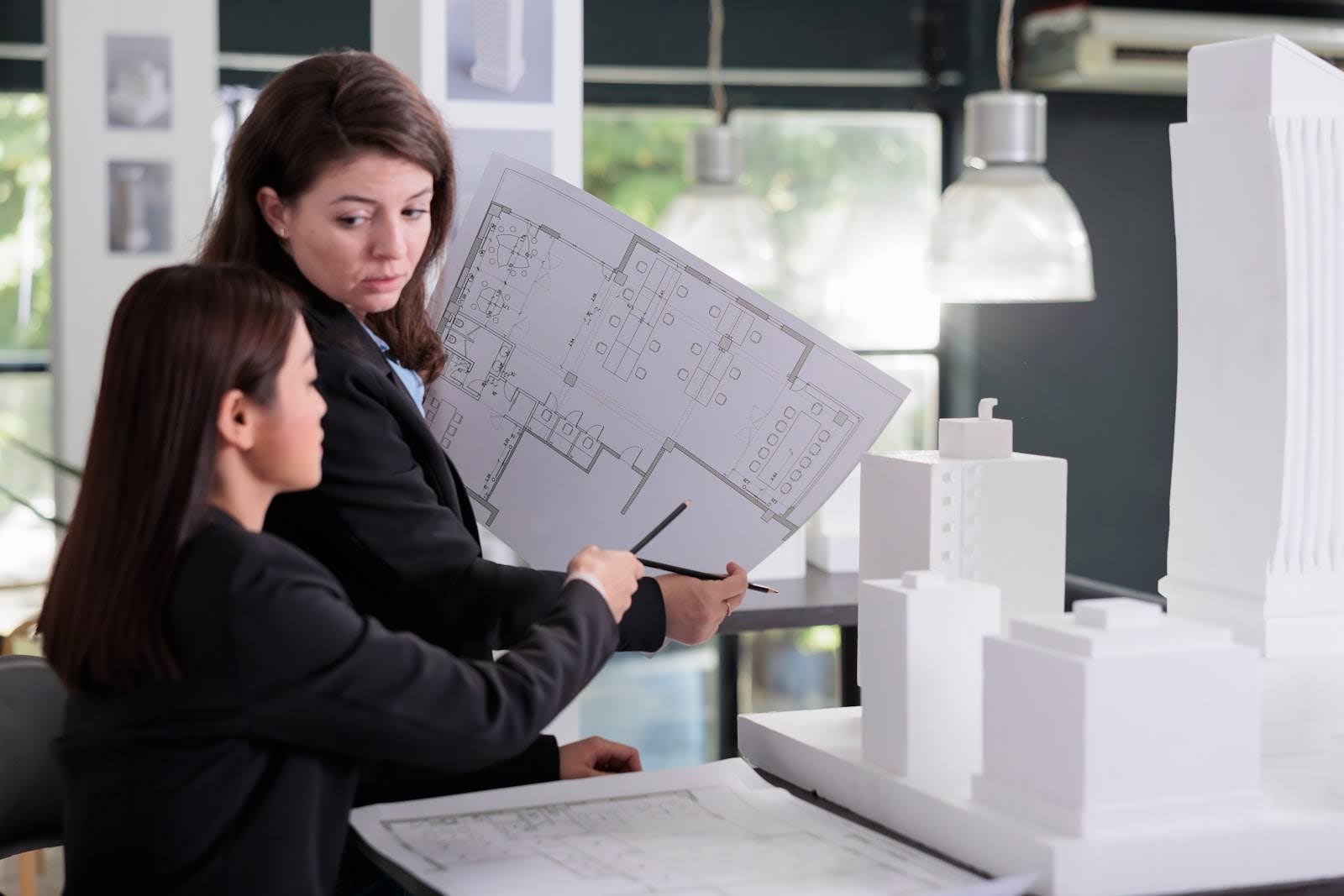 Two women in a modern office review architectural blueprints, focusing on resource capacity planning. One holds the plans while the other points at them. In the foreground, there are white architectural models of buildings, and bright windows illuminate the background.