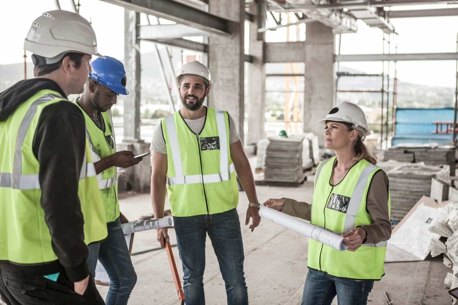 A group of five construction workers in safety vests and helmets stand on a building site, holding blueprints and discussing the billable vs non billable hours. Scaffolding and building materials are visible in the background.