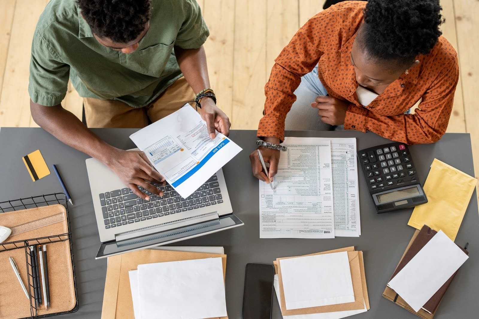 Two people sit at a desk filled with documents, a laptop, calculator, and stationery. They are working on paper forms, one holding a pen and the other examining a sheet. The scene suggests a focus on tax compliance and financial or administrative tasks.