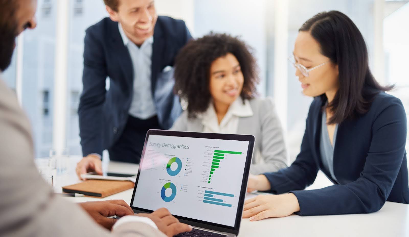 A diverse group of four people in business attire is gathered around a table, discussing a resource management system. One person uses a laptop displaying charts, including pie and bar graphs labeled "Survey Demographics." They are engaged in discussion and smiling.