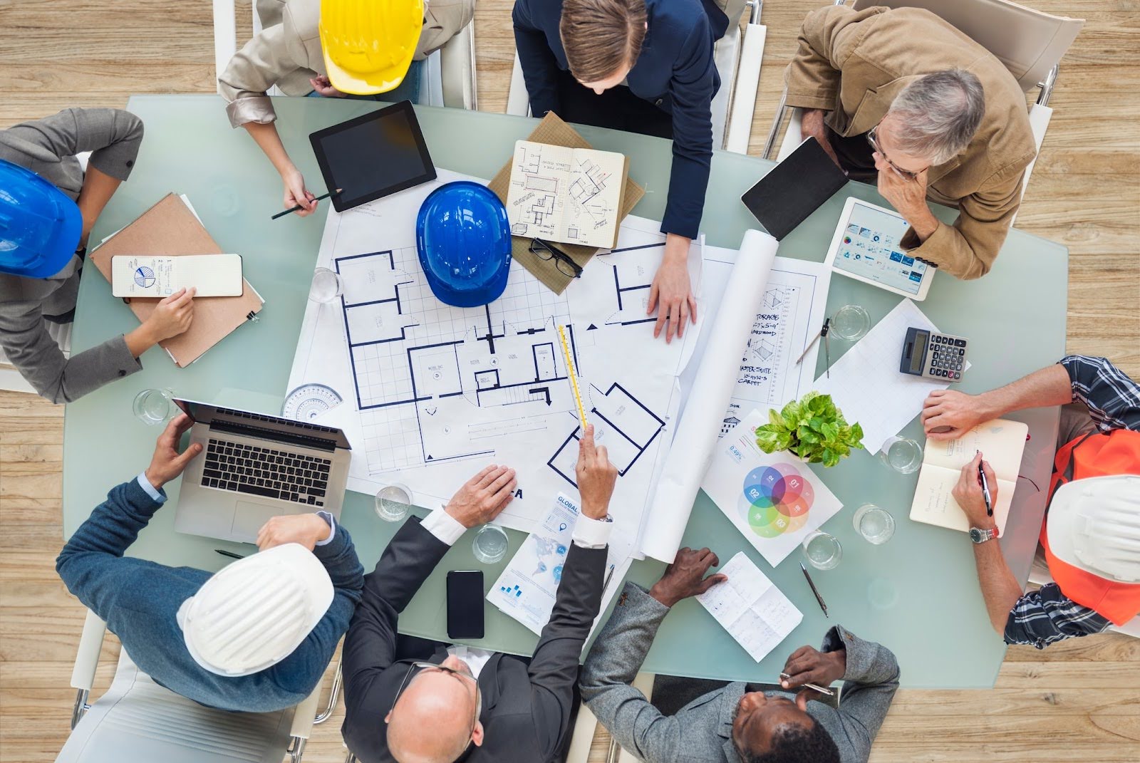 A group of diverse people in business attire and hard hats gather around a table with architectural blueprints, utilizing laptops, tablets, and notepads. The scene suggests a collaborative planning session enhanced by Deltek Vantagepoint for seamless asset management.