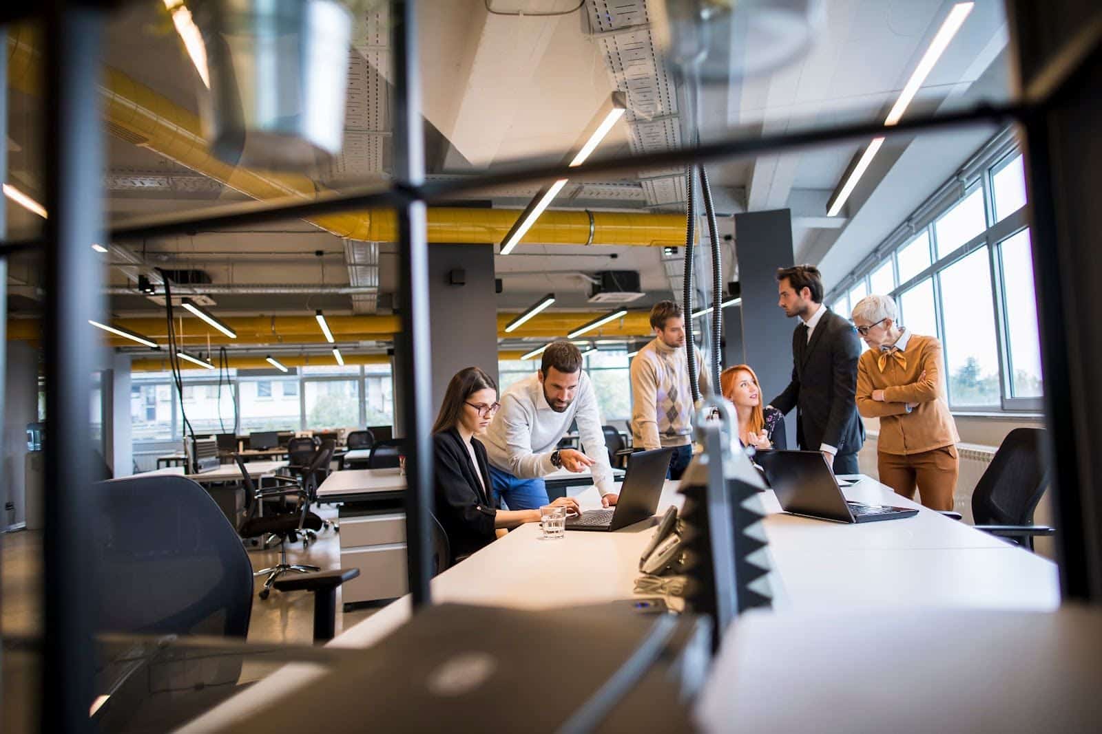 A group of six people gathered around a table in a modern open office. They are discussing something while looking at a laptop. The room has large windows, overhead lighting, and various office equipment.