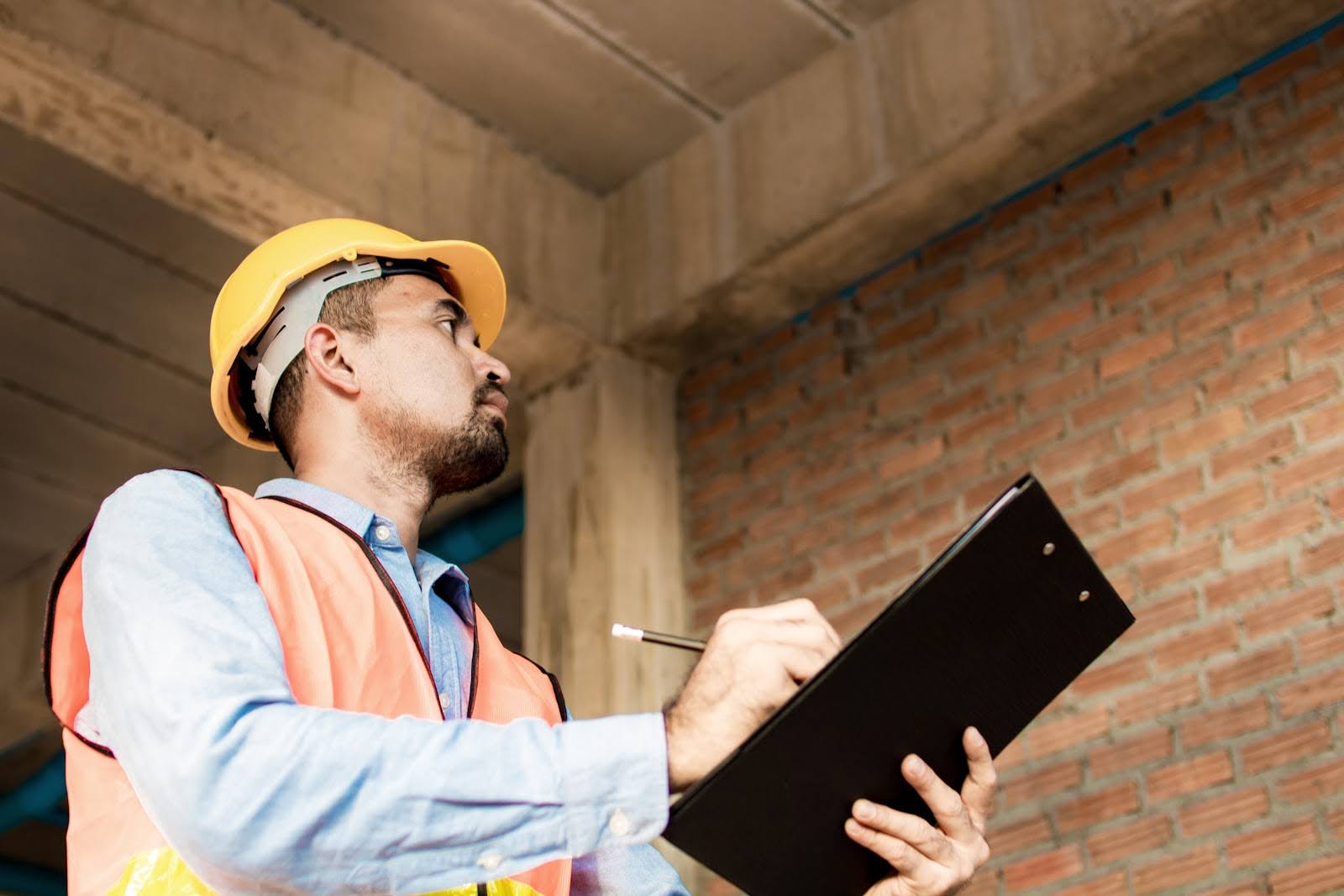 A construction worker wearing a yellow hard hat and orange vest holds a clipboard and pen, inspecting a brick wall inside an unfinished building while reviewing building commissioning software.