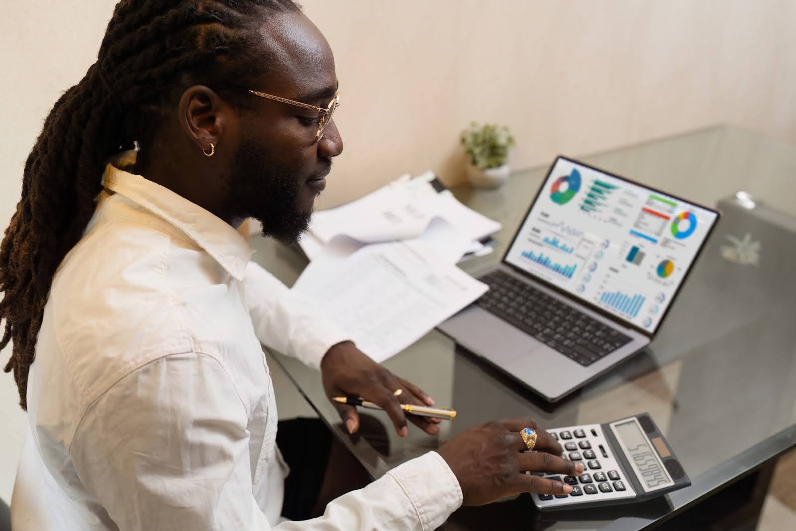 A person with long hair and glasses sits at a glass desk, using a calculator and reviewing printed documents. Nearby, a laptop with cash flow forecasting software displays colorful charts and graphs. A small plant is in the background.