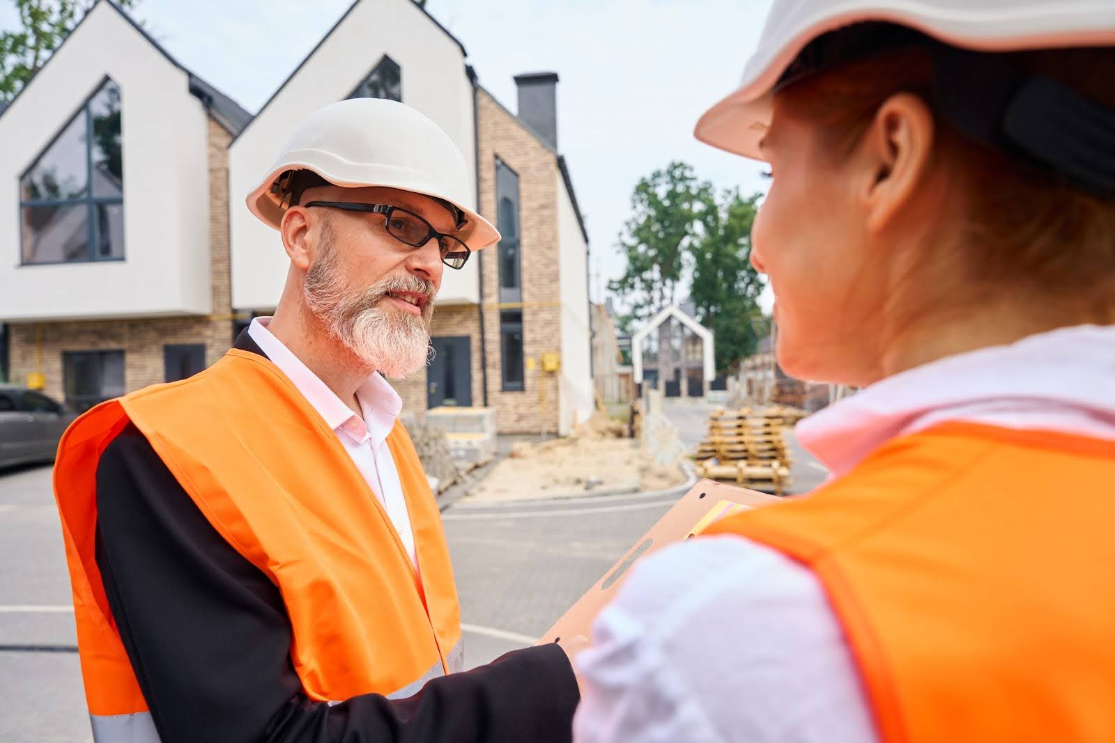 Two construction workers wearing orange vests and white hard hats are having a discussion at a construction site with modern houses in the background. They appear to be reviewing a clipboard. Trees and building materials are visible nearby.
