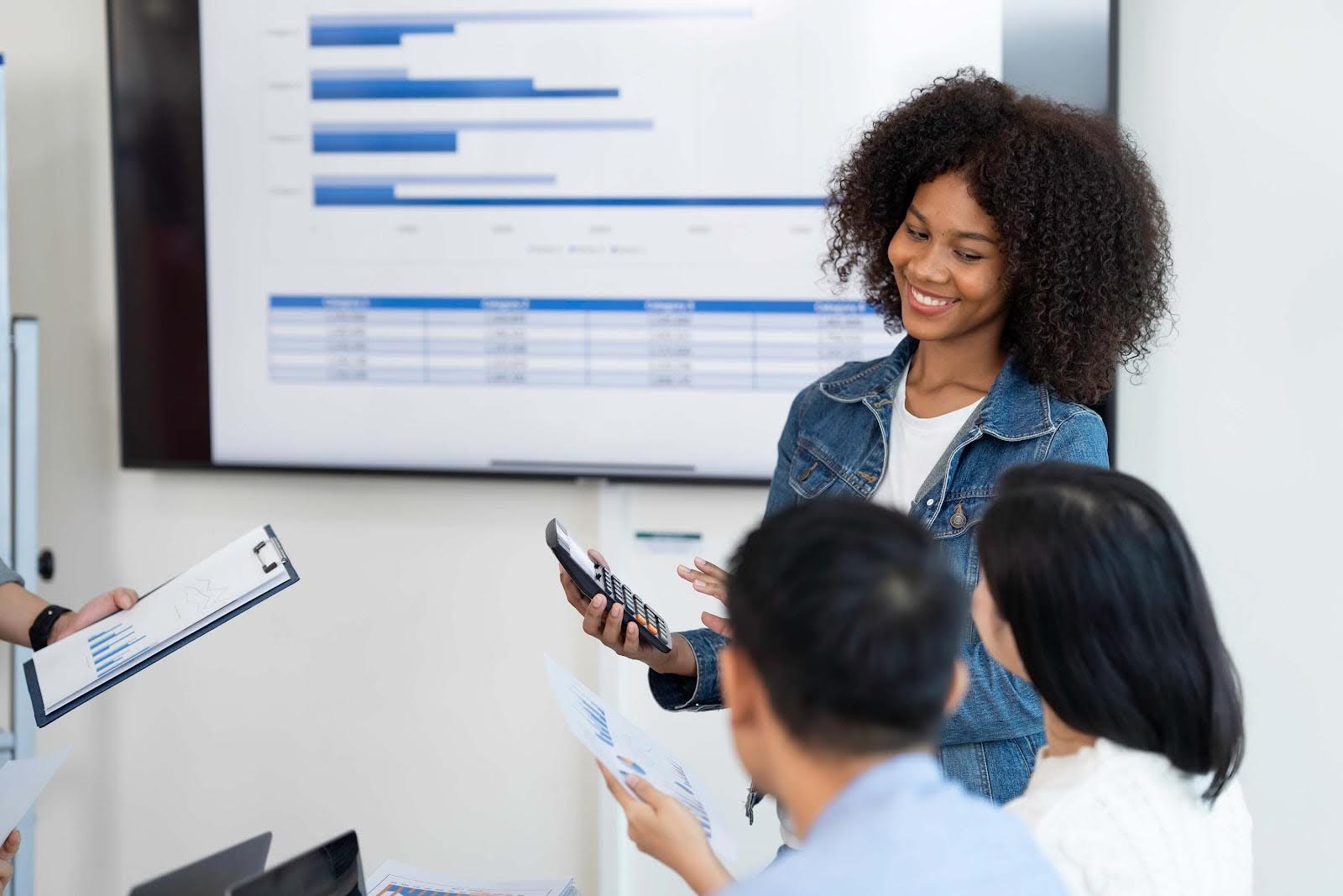 A woman smiles while presenting rolling forecasts to colleagues in a meeting room. She holds a calculator and a document, while others sit listening. A large screen behind displays charts and graphs.