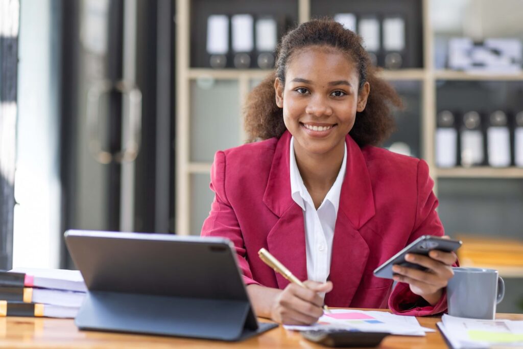 A young woman in a red blazer sits at a desk, smiling while holding a smartphone and writing on paper—balancing tasks with ease, just like managing accounting for professional services. A tablet, notebooks, and coffee mug are nearby; shelves and documents fill the background.