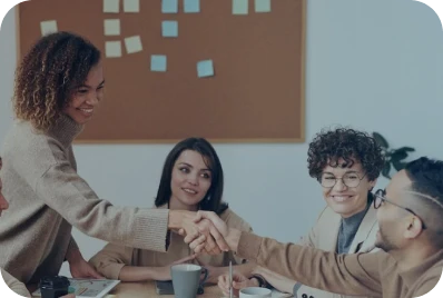 Four people sit around a table in a meeting room. Two of them, a woman and a man, are shaking hands and smiling. A corkboard with sticky notes is visible on the wall behind them.