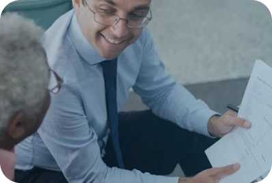 A man in glasses and a blue shirt is smiling and holding documents, showing them to an older person with gray hair during a discussion.