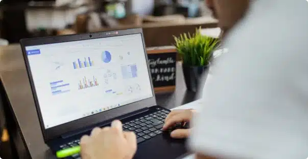 A person using a laptop displaying various charts and graphs, working at a desk with a small potted plant and a chalkboard sign in the background.