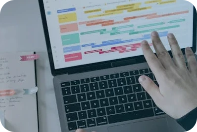 A person’s hand hovers over a laptop keyboard, which displays a colorful, detailed calendar or scheduling app. A notebook with handwritten notes and a pen are visible to the side on the desk.