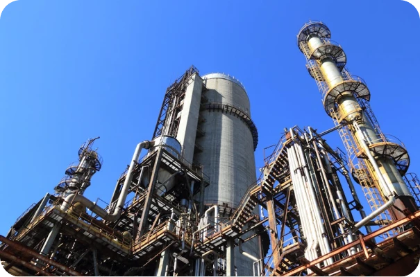Industrial refinery with several tall metal structures and towers against a clear blue sky. The complex pipes, platforms, and cylindrical units are part of a large-scale processing plant.