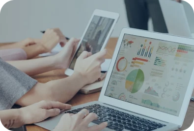 People working at a desk with a laptop displaying colorful charts and graphs, and another person using a tablet; hands are visible, suggesting a collaborative work or business meeting environment.