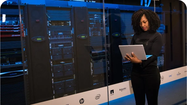 A woman stands in front of server racks in a data center, holding and working on a laptop. She is wearing a black outfit and appears focused on her work.