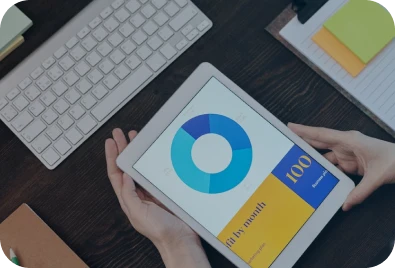 A person holding a tablet displaying a blue circular chart and the number 100, next to a keyboard, notepad, and colorful sticky notes on a dark desk.