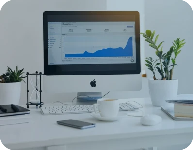 A modern desk with an iMac displaying a blue line graph, surrounded by potted plants, a cup of coffee, a keyboard, a mouse, a notebook, a smartphone, and an hourglass.