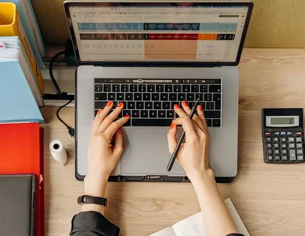 A person with red-painted nails types on a laptop keyboard while holding a stylus. The desk has a calculator, notebook, pen, and folders, indicating a work or study setting.