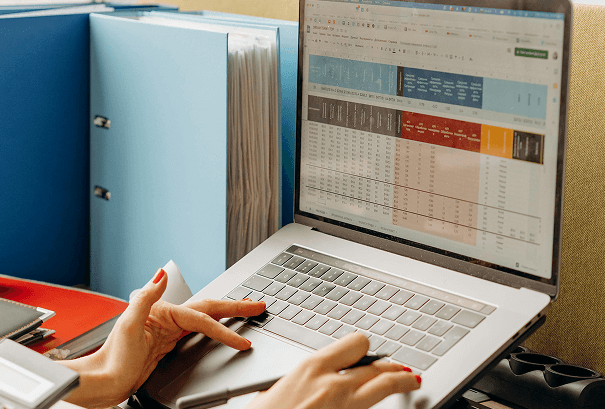 A person working on a laptop with a spreadsheet open on the screen, sitting next to a large blue file folder filled with documents. One hand is on the keyboard, the other holding a pen.