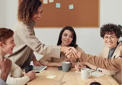 Four people sit around a table in an office, while two of them are shaking hands and smiling. Coffee cups, papers, and pens are on the table, and a corkboard with sticky notes is on the wall behind them.