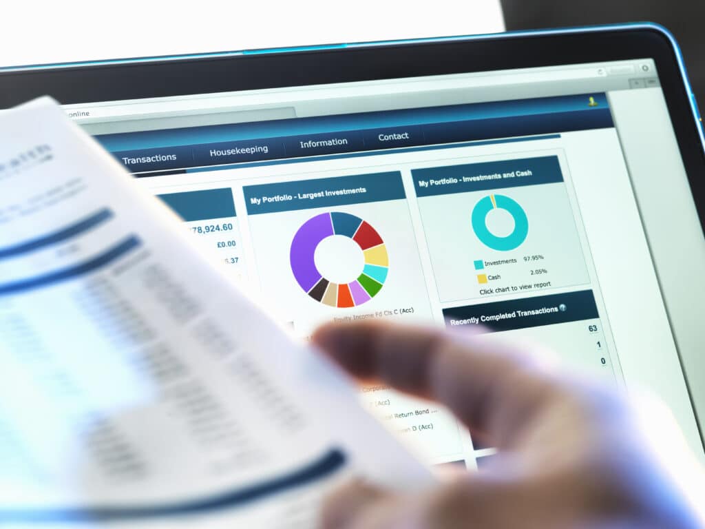 A person holds a financial document in front of a computer screen displaying colorful investment portfolio charts, graphs, and financial data on a dashboard interface.