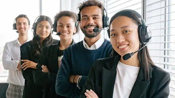 Five diverse customer service representatives wearing headsets stand in a row, smiling confidently in a modern office with large windows and blinds.