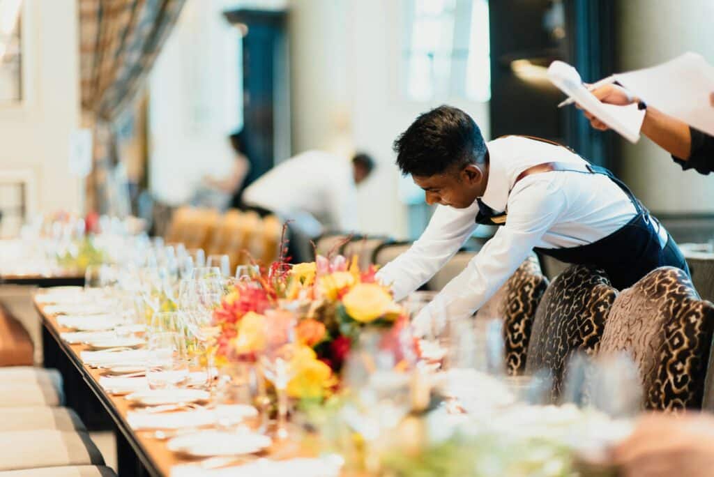 A server in a white shirt and black apron arranges a long, elegant dining table decorated with colorful flowers, glassware, and plates in a bright, upscale restaurant setting.