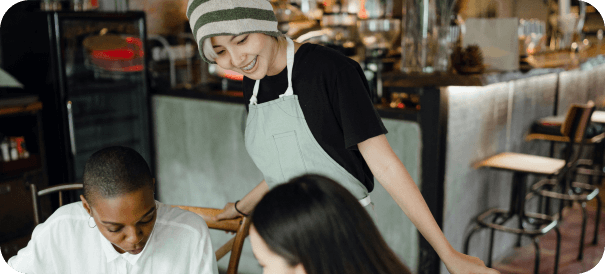 A smiling server wearing an apron and beanie stands by a table, interacting with two seated customers in a cozy café setting.