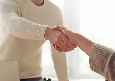 Two people wearing sweaters are shaking hands indoors, suggesting a greeting or agreement. Their faces are not visible.