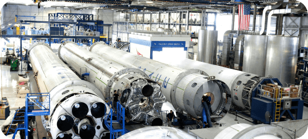 Large rockets are assembled inside a spacious, well-lit industrial facility, with numerous metal tanks, machinery, and an American flag visible in the background.