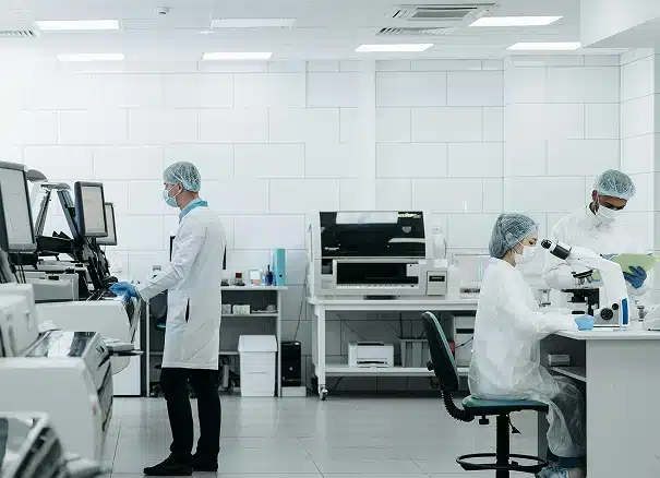 Three scientists in lab coats, masks, and hair covers work in a modern laboratory with lab equipment; one uses a computer, another looks in a microscope, and the third examines documents.