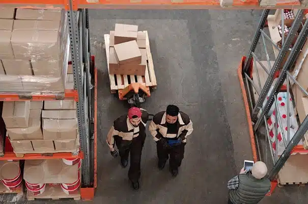 Overhead view of two warehouse workers in brown uniforms walking between shelves with boxes, a pallet jack carrying boxes between them, and another person nearby using a tablet.