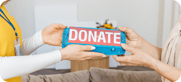Two people passing a blue donation box labeled "DONATE" in large letters, suggesting an act of giving or charity in a home or indoor setting.