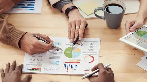 Four people sit at a table reviewing colorful charts and graphs on paper. One person points at a pie chart; others hold pens. A tablet, coffee cup, and more documents are also on the table.