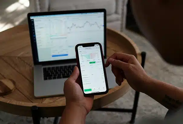 A person holds a smartphone displaying a stock trading app, while a laptop with financial charts open sits on a round wooden table in the background.