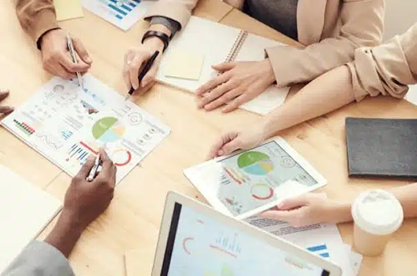 Four people sit at a wooden table reviewing charts and graphs on paper and a tablet. A laptop, notebook, and coffee cup are also on the table, suggesting a collaborative business meeting.