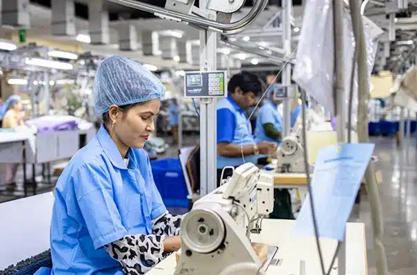 A woman wearing a blue hairnet and uniform operates a sewing machine in a garment factory, with other workers and machinery visible in the background.