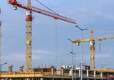 Several tall construction cranes stand above a building under construction, with scaffolding and unfinished concrete structures visible against a blue sky.