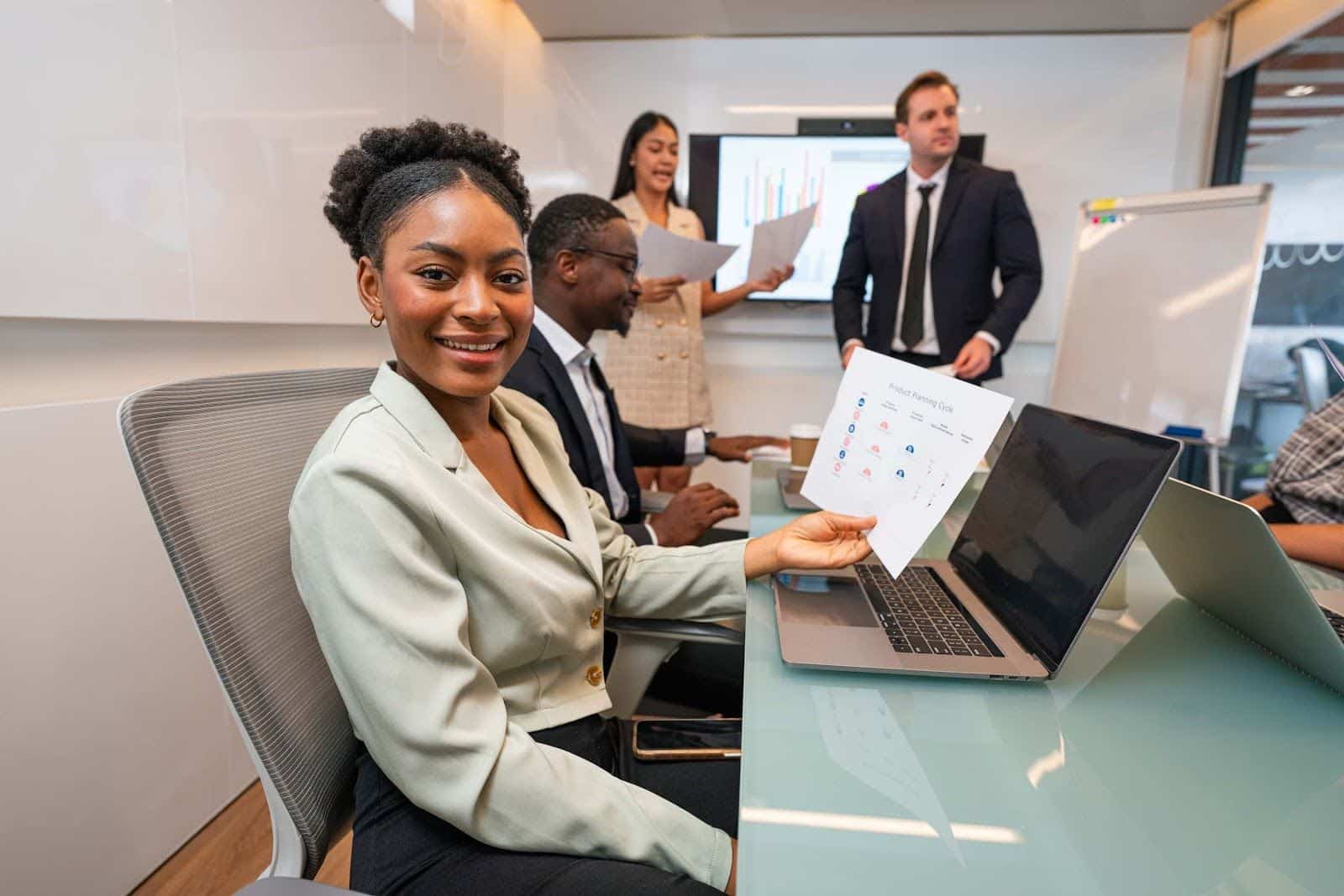 A woman smiles at the camera while holding a paper with charts in a meeting room. Three colleagues are in the background, discussing Sage professional services and documents near a laptop and a whiteboard, showcasing teamwork in professional services.