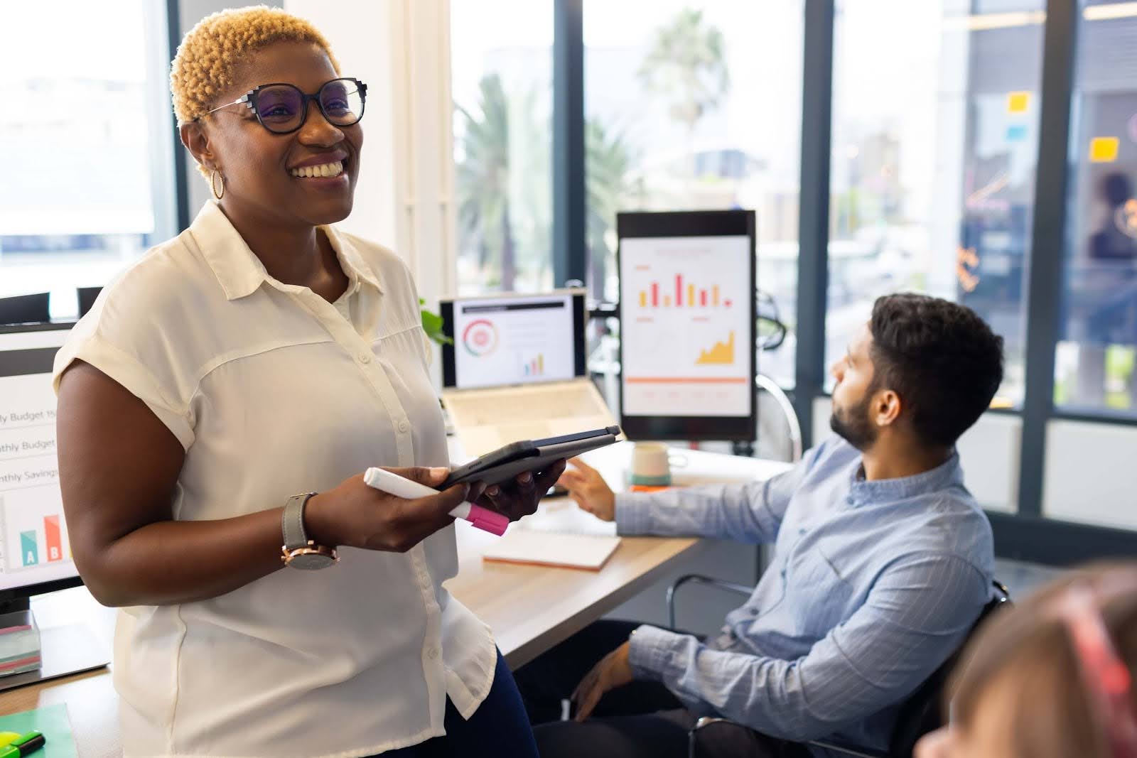 A smiling woman holding a tablet stands in an office, while a man reviews financial data on computer monitors. Sunlight streams through large windows in the modern workspace, reflecting the efficiency of sage intacct financial services.