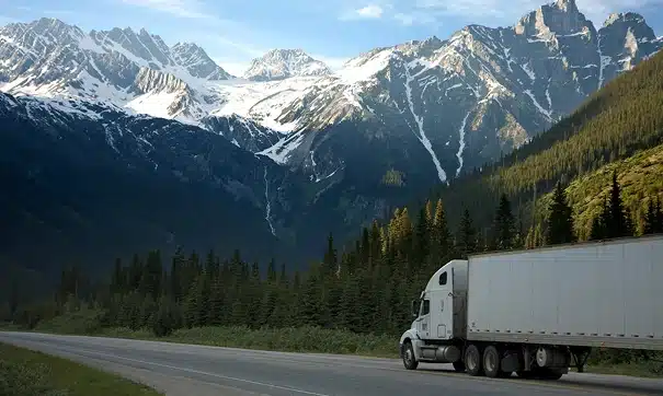 A white semi-truck drives along a highway surrounded by dense pine forests, with tall, snow-capped mountains rising in the background under a blue sky.