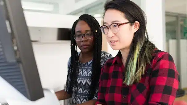 Two women are working together at a computer in an office. One wears glasses and a red plaid shirt, while the other has long braids and glasses. Both are focused on the monitor.