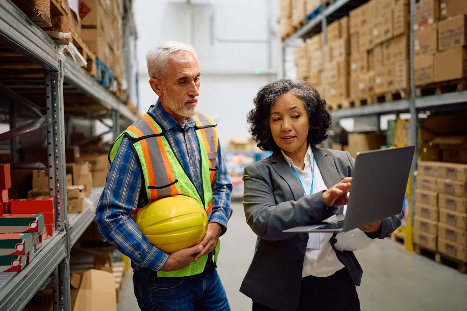 Two workers in a warehouse discuss something on a laptop. One, in a suit, points at the screen while the other, in a safety vest and holding a yellow hard hat, listens intently—perhaps reviewing accounting processes for their manufacturing business.