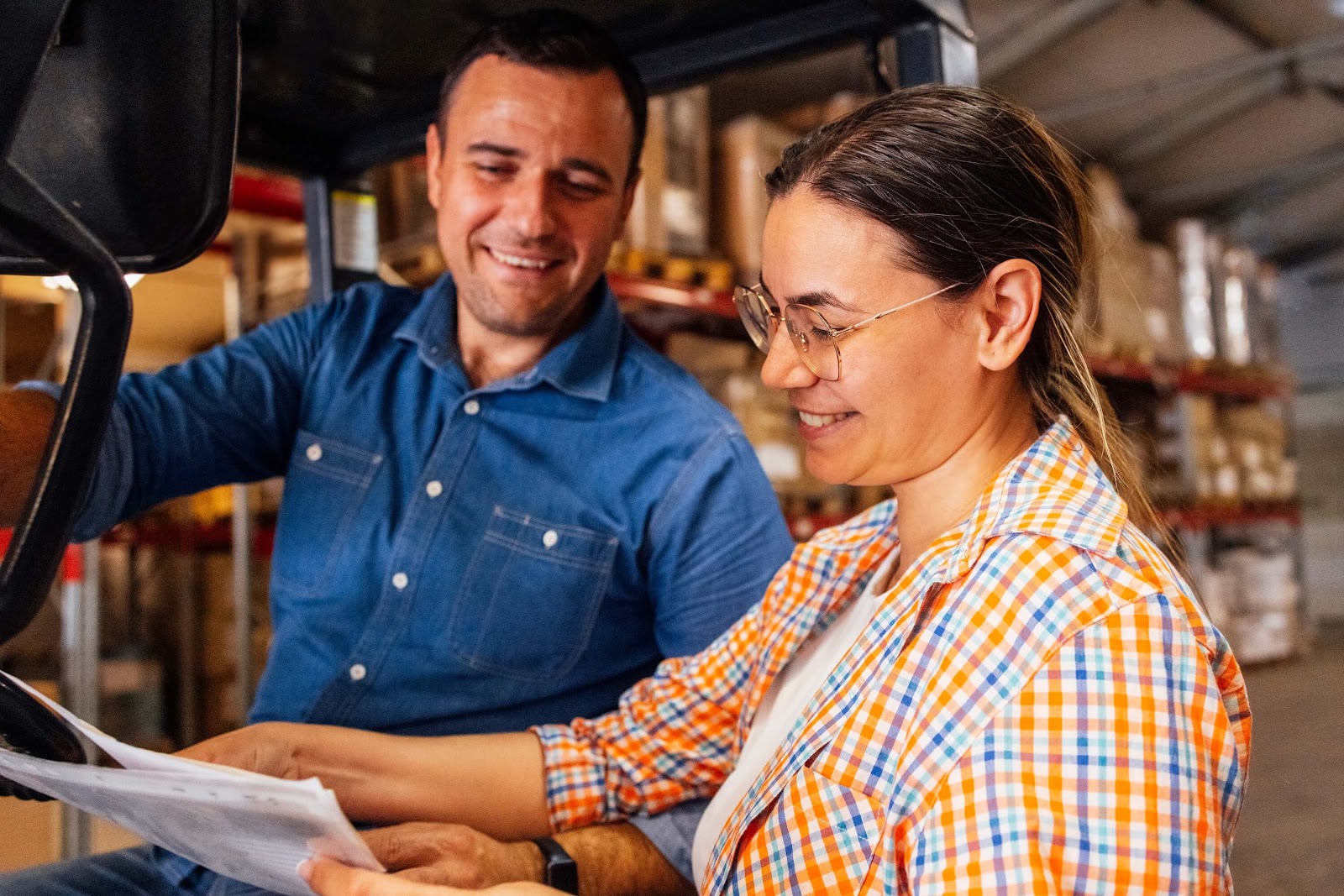 Two people in a warehouse, surrounded by shelves of boxes, smile and review documents together. Seated and standing, they appear engaged, collaborating on accounting processes for their manufacturing business.