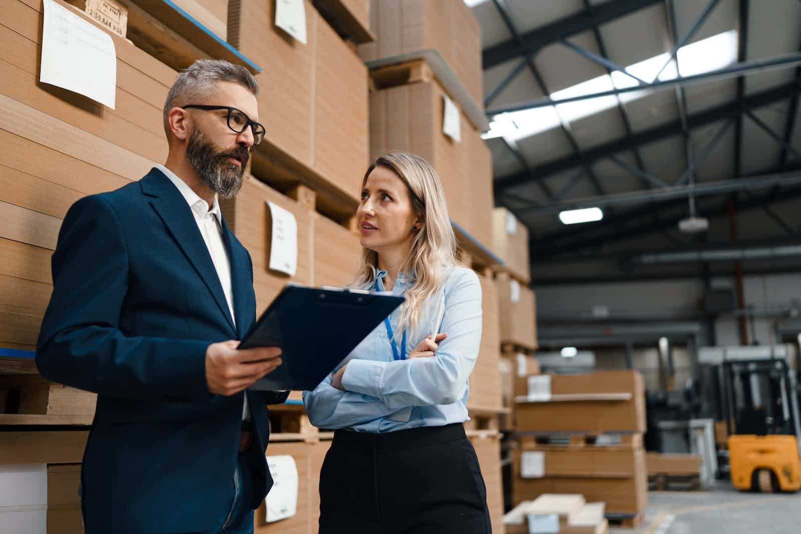 A man in a suit holding a clipboard discusses manufacturing accounting challenges with a woman in a warehouse filled with stacked cardboard boxes and shelves. The organized, industrial setting emphasizes the demands of managing a manufacturing business.